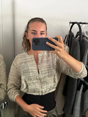 Beige croppad overshirt med dragkedja - Säljer en beige croppad overshirt med diskret rutigt mönster och dragkedja framtill. Jackan har lång ärm, elastisk midja och är tillverkad i ett lätt, strukturerat material som känns luftigt. Perfekt för lager på lager och en avslappnad stil.