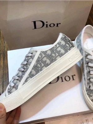 Grå Dior sneakers med monogram - Snygga sneakers från Dior i grått och vitt med det klassiska Dior-monogrammet över hela skon. Skorna har vita snören, metallfärgade öljetter och en vit sula med 'Christian Dior' präglat på sidan. Perfekta för dig som vill ha en exklusiv och trendig look.