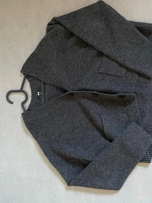 Mörkgrå stickad tröja med knappar - Mysig mörkgrå stickad tröja med knappar framtill och två fickor. Tröjan har ribbstickade muddar vid ärmslut och nederkant samt en bred krage. Perfekt för kyliga dagar och enkel att matcha med olika outfits.