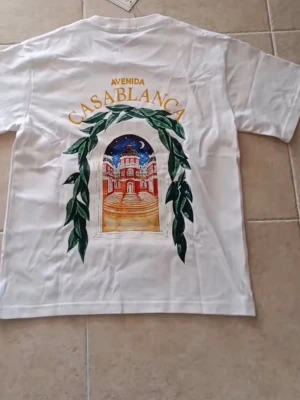 Vit t-shirt Casablanca Avenida - Vit t-shirt från Casablanca med färgglad tryck på både bröstet och ryggen. Motivet visar en byggnad med gröna blad och texten 'Avenida Casablanca' i gult. Klassisk passform och rund hals. Perfekt för dig som gillar stilrena men ändå unika plagg.