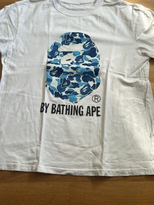 Vit BAPE t-shirt med blått camomönster - Säljer en vit t-shirt från By Bathing Ape med det ikoniska apansiktet i blått camomönster på bröstet och texten 'BY BATHING APE' under. Klassisk rund hals och korta ärmar. Perfekt för dig som gillar streetwear och vill sticka ut.
