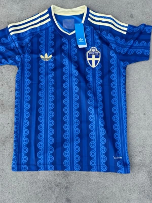 Sverige Borta VM tröja 2026 - Retroinspirerad blå fotbollströja från Adidas med Sveriges landslagsemblem på bröstet. Tröjan har ljusgula detaljer, klassiska Adidas-ränder på axlarna och ett mönster i olika blå nyanser. Tillverkad i lätt och ventilerande material. Strlk S