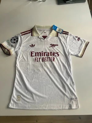 Helt ny! Arsenal 25/26 tredje Gyökeres  - Snygg vit Arsenal bortatröja från Adidas med vinröda detaljer och klubbmärke på bröstet. Tröjan har korta ärmar, krage och tryckta sponsorer samt Champions League-märke på ärmen. Namnet Gyökeres och nummer 14 på ryggen. Tillverkad i lätt funktionsmaterial.