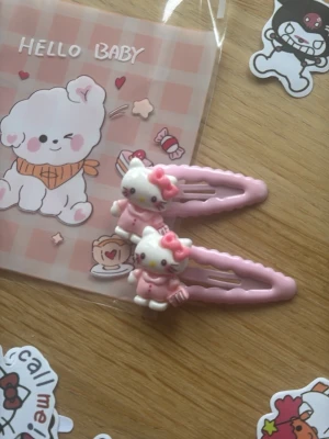 Söta rosa Hello Kitty hårspännen - Två gulliga hårspännen i ljusrosa metall med 3D Hello Kitty-figurer i rosa klänning och rosett. Perfekta för att piffa upp frisyren med en söt och lekfull touch. Passar dig som gillar kawaii och detaljerade accessoarer.
