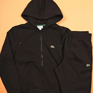 Lacoste tracksuit - Säljer en svart hoodie från Lacoste med dragkedja framtill och klassisk huva med dragsko. Hoodien har det ikoniska krokodilmärket broderat på bröstet och en stor ficka framtill. Perfekt för en avslappnad och sportig stil.