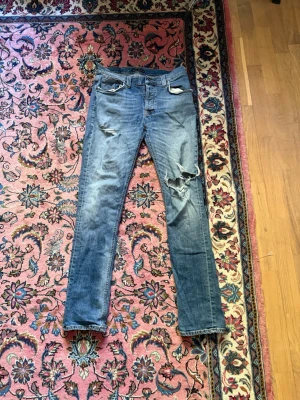 Slitna Nudie Grim Tim - Säljer ett par blå skinny jeans med slitningar och hål på låret och knät. Jeansen har klassisk femficksdesign och är tillverkade i denim med stretch. Perfekta för en avslappnad och trendig look. W34 L34