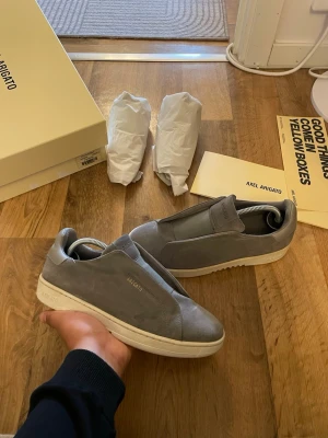 Grå sneakers från  Axel Arigato - Snygga grå slip-on sneakers från Axel Arigato i mocka med diskret logga i guld på sidan. Skorna har en vit, chunky sula och minimalistisk design utan snörning. Perfekta för dig som gillar stilrena och moderna sneakers. Storlek 40 passar 41