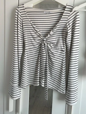 Randig långärmad topp med snörning - Säljer en vit och svart randig topp från Design by Si. Toppen har långa ärmar, djup v-ringning och snygg snörning framtill som ger en trendig look. Perfekt för dig som gillar stilrena plagg med en twist.