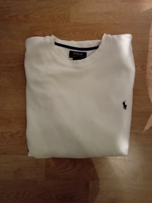 Vit långärmad tröja från Polo Ralph Lauren -  stilren vit långärmad tröja från Polo Ralph Lauren. Tröjan har rund halsringning och den klassiska broderade loggan i marinblått på bröstet. Perfekt basplagg för en clean look. Säljes då den ej passar min stil längre använd 1 gång i min ägo, Tveka inte på att höra av er :)
