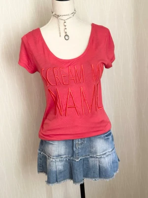 Rosa t-shirt med text och öppen rygg - Trendig rosa t-shirt med djup urringning fram och bak samt cool texten 'SCREAM MY NAME' broderad på framsidan. T-shirten har korta ärmar och en öppen ryggdetalj med band. Perfekt för dig som vill sticka ut med en unik och ungdomlig stil.