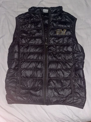 Svart dunväst från EA7 Emporio Armani - Snygg svart dunväst från EA7 Emporio Armani med quiltad design och dragkedja framtill. Västen har en hög krage och logga i guld på bröstet. Perfekt att slänga över en hoodie eller tröja för extra värme och stil.