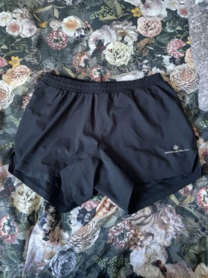 Svarta träningsshorts från Ronhill - Säljer ett par svarta träningsshorts från Ronhill med elastisk midja och kort passform. Lätta och luftiga, perfekta för löpning eller gym. Diskret logga på ena benet och slits i sidan för extra rörelsefrihet.