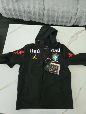 Svart Brasil vindjacka med huva - Snygg svart vindjacka med huva och dragkedja, prydd med Brasil-logga, gula och röda detaljer samt flera tryck som Itaú och vivo. Jackan är tillverkad i slitstarkt GORE-TEX-material och har fickor med dragkedja. Perfekt för dig som gillar sportig stil.