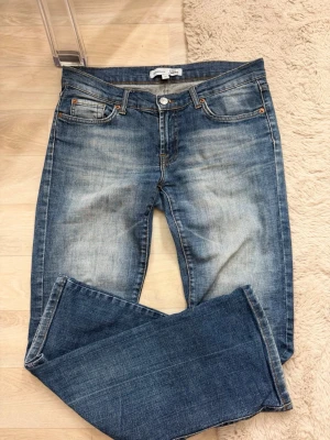 Blå bootcut jeans med slitningar - Säljer ett par blå bootcut jeans med snygga slitningar och tvättade detaljer. Jeansen har klassisk femficksmodell, normal midja och dragkedja med knapp. Perfekta för en avslappnad och trendig look. En flärp på sidan av jeansen har lossnat men går säkert att sy tillbaka