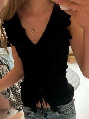 Svart v-ringad blus med snörning - Snygg svart blus med v-ringning och volangdetaljer vid axlarna. Toppen är ärmlös och har snörning framtill som ger en trendig look. Perfekt att styla med jeans eller kjol för en cool outfit.