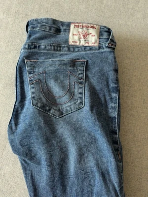  True Religion jeans - Säljer ett par klassiska blå jeans från True Religion med ikoniska kontrastsömmar och stora bakfickor med broderad logga. Jeansen har normal passform och är tillverkade i mjukt denimtyg. Perfekta för en avslappnad och trendig look.