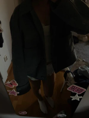 Mörkgrå cardiganjacka med stjärnlapp - Säljer en mörkgrå cardiganjacka med rak passform och krage. Jackan har en cool detalj med en rödvit stjärnlapp på ena ärmen. Perfekt för dig som vill ha en enkel men unik jacka till din outfit.