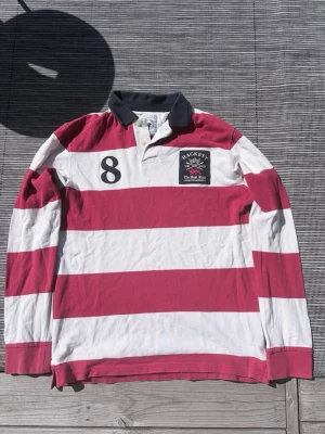 Randig rugbytröja från Hackett London - Säljer en långärmad rugbytröja från Hackett London i röd och vit med breda ränder. Klassisk krage i mörkblått, broderad logga och siffran 8 på bröstet. Tillverkad i bomull, perfekt för en sportig och avslappnad stil.