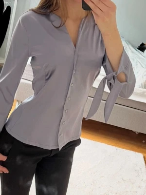 Blå satinblus med knytband - Snygg blå satinblus med knappar framtill och långa ärmar med dekorativa knytband vid handlederna. Blusen har en lätt v-ringad halsringning och en figurnära passform som ger en elegant look. Perfekt att styla med jeans eller kjol för en trendig outfit.