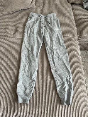 Gråa mjukisbyxor från Nike - Säljer ett par gråa mjukisbyxor från Nike med dragsko i midjan och ribbade muddar vid bensluten. Byxorna har en klassisk passform och Nike-logga på vänster lår. Perfekta för chill dagar eller träning.