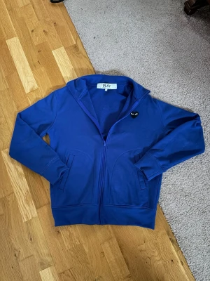 Blå CDG Play Zip Hoodie - Säljer en blå hoodie med dragkedja och huva från PLAY. På bröstet sitter en liten svart hjärtdetalj med ögon och på ryggen finns ett stort svart hjärta med ögon. Hoodien har fickor på sidorna och är i mjukt material, perfekt för en chill och stilren look.