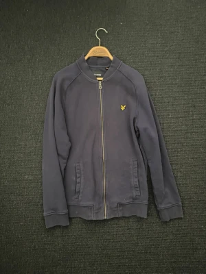 Mörkblå ziptröja från Lyle & Scott - Mörkblå ziptröja från Lyle & Scott med gul logga på bröstet. Tröjan har ribbade muddar, två fickor framtill och en klassisk bomberkrage. Perfekt för en avslappnad och stilren look.