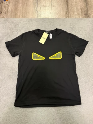 Svart Fendi t-shirt med gula ögon - Svart t-shirt från Fendi med korta ärmar och ett par gula ögon i grafiskt tryck på bröstet. T-shirten har en klassisk passform och är tillverkad i mjuk bomull. Perfekt för dig som vill sticka ut med en cool detalj.