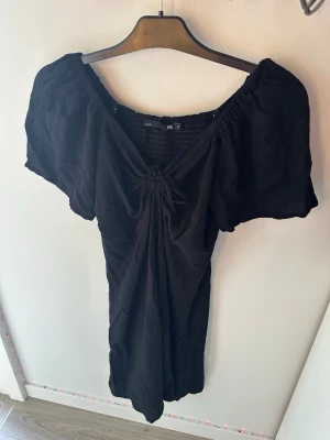 Svart off shoulder klänning 157 - Svart off shoulder klänning från 157 med korta puffärmar och smock upptill. Klänningen har en knytning framtill och är gjord i ett mjukt, tunt tyg som känns skönt mot huden. Perfekt för en trendig och avslappnad look.