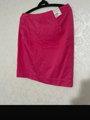 Rosa kortkjol med dragkedja - En snygg rosa kortkjol med dekorativ rosett framtill och guldfärgad dragkedja bak. Kjolen har en stilren design och är perfekt för dig som gillar färg. Materialet känns slätt och lätt, vilket gör den bekväm att bära.