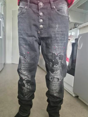 Svarta Philipp Plein jeans med dödskallar - Säljer ett par svarta jeans från Philipp Plein med tryckta dödskallar och färgstänk i rött och vitt. Jeansen har fem knappar framtill, smal passform och coola detaljer som små nitar vid fickorna. Perfekta för dig som vill sticka ut med din stil. Byxorna var lite nötta i grenen och har därför förstärkts med stödstygn från insidan. Syns ej när man har byxorna på sig. Taggen med storlek och material är bortklippt sedan tidigare. Storlek 30/32