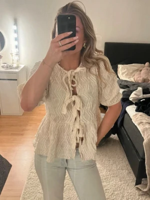 Krämvit topp med knytning fram - Trendig krämvit topp med korta puffärmar och snygg struktur i tyget. Toppen har flera knytningar framtill och en lätt utsvängd passform. Perfekt för dig som gillar en romantisk och bohemisk stil.