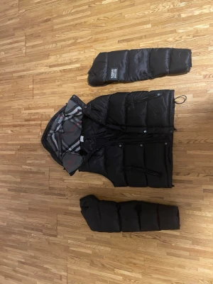 Svart burberry jacka med avtagbara ärmar - Svart bruberry jacka med avtagbara ärmar och huva. Jackan har rutigt innerfoder i grått, vitt och rött samt dragkedja och knappar framtill. Praktiska fickor på sidorna och justerbar nederkant. Perfekt för kalla vinterdagar.