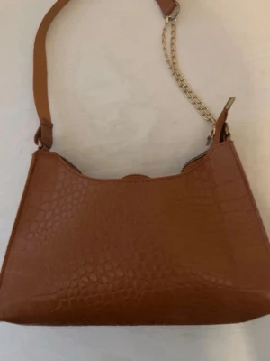 Brun axelremsväska med kedjedetalj - Snygg brun handväska i croco-mönstrat konstläder med axelrem och guldfärgad kedjedetalj. Väskan har en enkel, stilren design och stängs med dragkedja. Perfekt för dig som gillar trendiga accessoarer med lite edge.