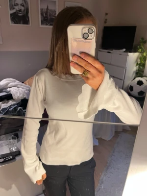 Vit långärmad topp med volangärm - Säljer en vit långärmad topp med ribbad struktur och unika volangdetaljer vid ärmslut och halsringning. Toppen har en slim passform och är tillverkad i ett mjukt, stretchigt material som känns bekvämt mot huden.