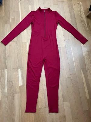 Röd figurnära jumpsuit med dragkedja – strl M - Snygg röd jumpsuit med långa ärmar och hög krage. Dragkedja framtill som går ner till midjan. Tight passform och långa ben. Materialet är stretchigt och bekvämt, perfekt för en trendig och sportig look.