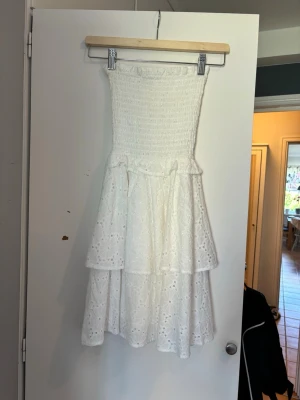 Vit axelbandslös volangklänning - Söt vit axelbandslös klänning med smock upptill och två lager volanger. Klänningen har broderade spetsdetaljer och ett knytband i midjan. Perfekt för sommarens alla tillfällen! Nästan aldrig använd