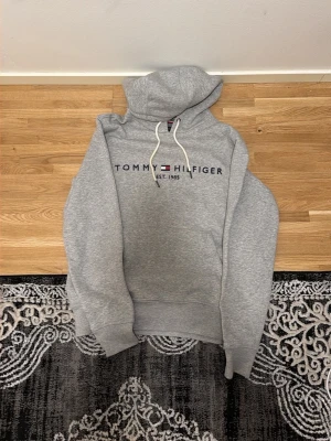 Grå hoodie från Tommy Hilfiger - Snygg grå hoodie från Tommy Hilfiger med broderad logga och text på bröstet. Klassisk huva med vita dragsnören och mjukt material som känns skönt mot huden. Perfekt för en avslappnad och stilren look.