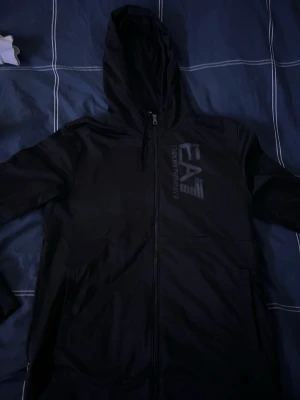 Armani tracksuit - Snygg svart hoodie och svarta mjukisbyxor  från Emporio Armani EA7 med dragkedja framtill och stor logga på bröstet. Tillverkad i mjukt material och har två fickor samt justerbar huva med snören. Perfekt för en chill och stilren look.    Fråga om ni har nån fundering  