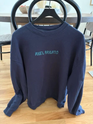 Mörkblå sweatshirt från Axel Arigato - Mörkblå sweatshirt med rund hals och tryckt Axel Arigato-logga i turkos framtill. Tröjan har avslappnad passform och ribbade muddar vid ärmslut och nederkant. Perfekt för en chill och stilren look.
