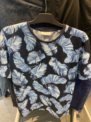 Blå mönstrad t-shirt från Lindbergh - Snygg t-shirt från Lindbergh med blått blad-mönster på mörkblå botten. T-shirten har rund halsringning och korta ärmar. Perfekt för dig som vill sticka ut med en cool och fräsch look.