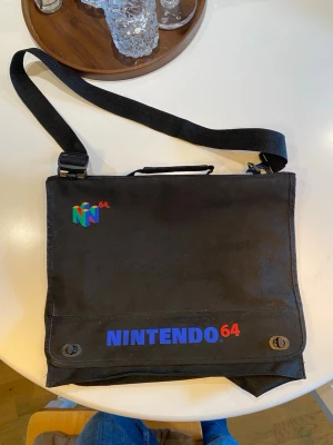 Nintendo 64 datorväska svart - Säljer en svart datorväska med Nintendo 64-logga i färg och texten NINTENDO 64 i blått och rött på framsidan. Väskan har justerbar axelrem och handtag, samt två spännen framtill. Perfekt för dig som gillar retro och gamingstil.