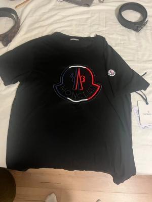 Svart Moncler t-shirt med stor logga - Säljer en svart t-shirt från Moncler med en stor, broderad logga i rött, blått och vitt på bröstet. T-shirten har korta ärmar och en klassisk passform. Liten Moncler-patch på ena ärmen. Perfekt för dig som gillar streetwear och exklusiva märken.