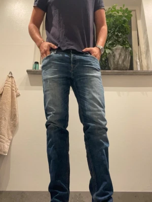 Blå raka jeans med slitningar - Säljer ett par klassiska blå jeans med raka ben och snygga slitningar. Jeansen har fem fickor och normal midja. Perfekta för en avslappnad och trendig look. Materialet är denim i bomull och färgen är mellanblå med ljusare partier.