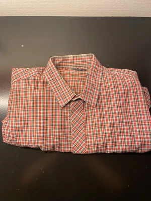 Röd och grön rutig skjorta - Säljer en klassisk rutig skjorta i rött, grönt och vitt. Skjortan har en traditionell krage och knäppning framtill. Perfekt för dig som gillar tidlös stil och vill ha något som sticker ut lite extra. Materialet känns lätt och bekvämt.