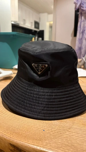 Prada hat - Snygg svart bucket hat från Prada med det ikoniska metalltriangel-logotypen framtill. Hatten är tillverkad i ett slitstarkt, lätt material som ger en clean och modern look. Perfekt accessoar för att lyfta din outfit och ge en lyxig touch.