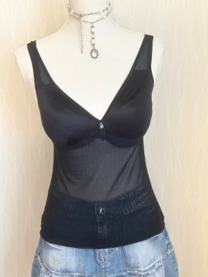 Svart mesh linnetopp med v-ringning - Snygg svart linnetopp i mesh med v-ringning och smala axelband. Toppen har en transparent, ribbad nederdel och en liten dekorativ detalj framtill. Perfekt att styla med jeans eller kjol för en trendig look.