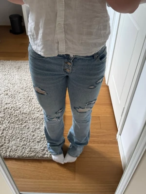 Ljusblå slitna bootcut jeans - Säljer ett par ljusblå jeans med slitningar på både fram- och baksida. Jeansen har bootcut-modell och knäppning med tre knappar framtill. Snyggt slitna detaljer ger en avslappnad och trendig look. 