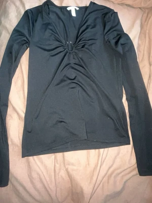Svart långärmad topp från H&M - Snygg svart långärmad topp från H&M med v-ringning och twistad detalj framtill. Tillverkad i ett stretchigt och mjukt material som sitter bekvämt på kroppen. Perfekt att styla med jeans eller kjol för en trendig look.