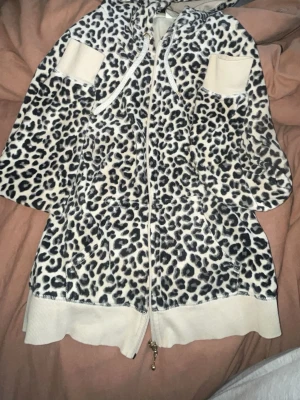 Leopardmönstrad hoodie med dragkedja - Säljer en snygg hoodie i leopardmönster med dragkedja framtill och huva med snören. Tröjan har ribbade muddar vid ärmslut och nederkant samt två fickor fram. Färgen är vit med svarta och grå fläckar. Perfekt för dig som gillar djurmönstrat och vill sticka ut.