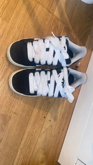 Adidas skor  - Säljer ett par svarta sneakers med breda vita skosnören och vit sula. Skorna har en låg profil och en klassisk siluett. Materialet ser ut att vara mocka eller liknande tyg och de har en vit logga på plösen.